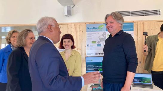 Photo of a group of individuals at a GreenDeal-NET event. On the right CIES-Iscte researchers Helge Jörgens and Inês Rocha Trindade. On the left Prime Minister António Costa. They are standing in front of a GreenDeal-NET promotional banner in a room with wooden paneling.